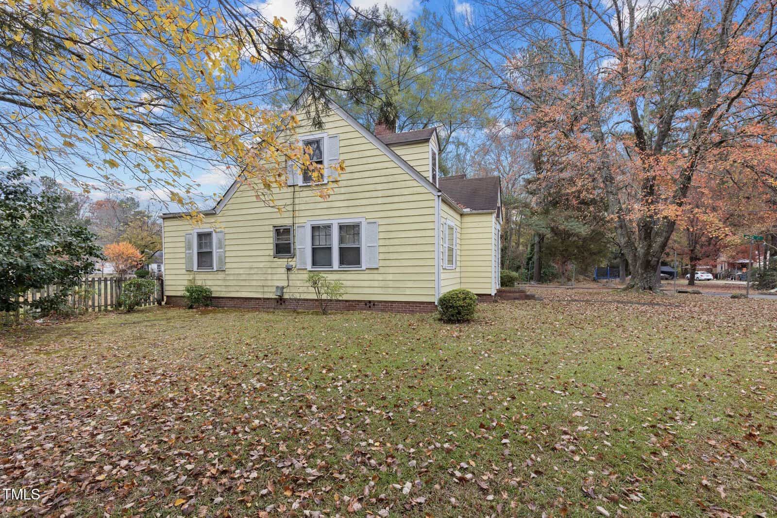 A yellow two-story house with white trim sits on a grassy yard scattered with autumn leaves. Several windows are visible on the side of the house, and trees with fall foliage surround the property.