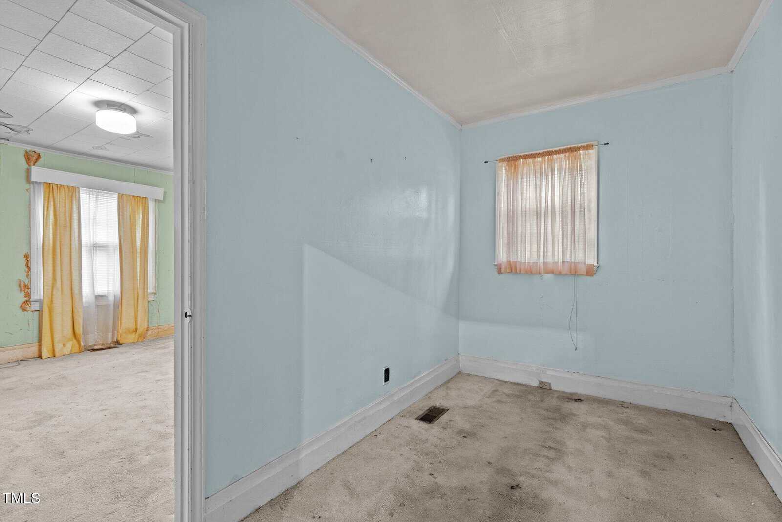 A small empty room with light blue walls, a single window with sheer peach curtains, and a beige carpet. An open doorway leads to a larger room with pale green walls and yellow curtains.