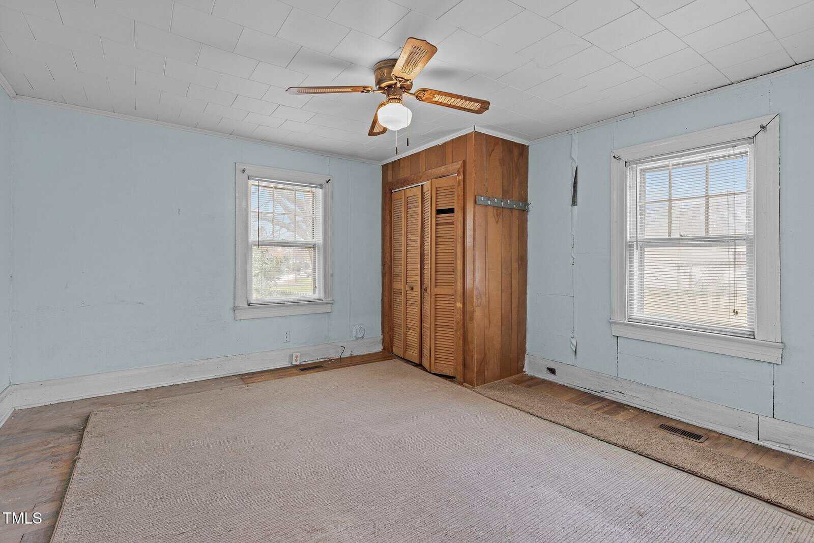 Empty room with light blue walls, two windows with blinds, a ceiling fan with light, a wooden closet with louvered doors, and a beige carpet covering part of the floor.