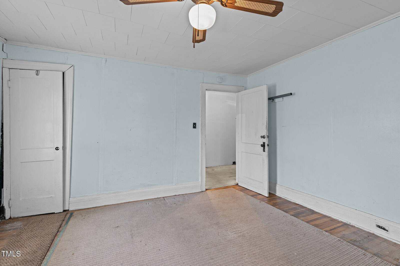 Empty room with light blue walls, white ceiling, open closet door, white door leading to another space, ceiling fan with light, and partially exposed hardwood flooring along the edge.