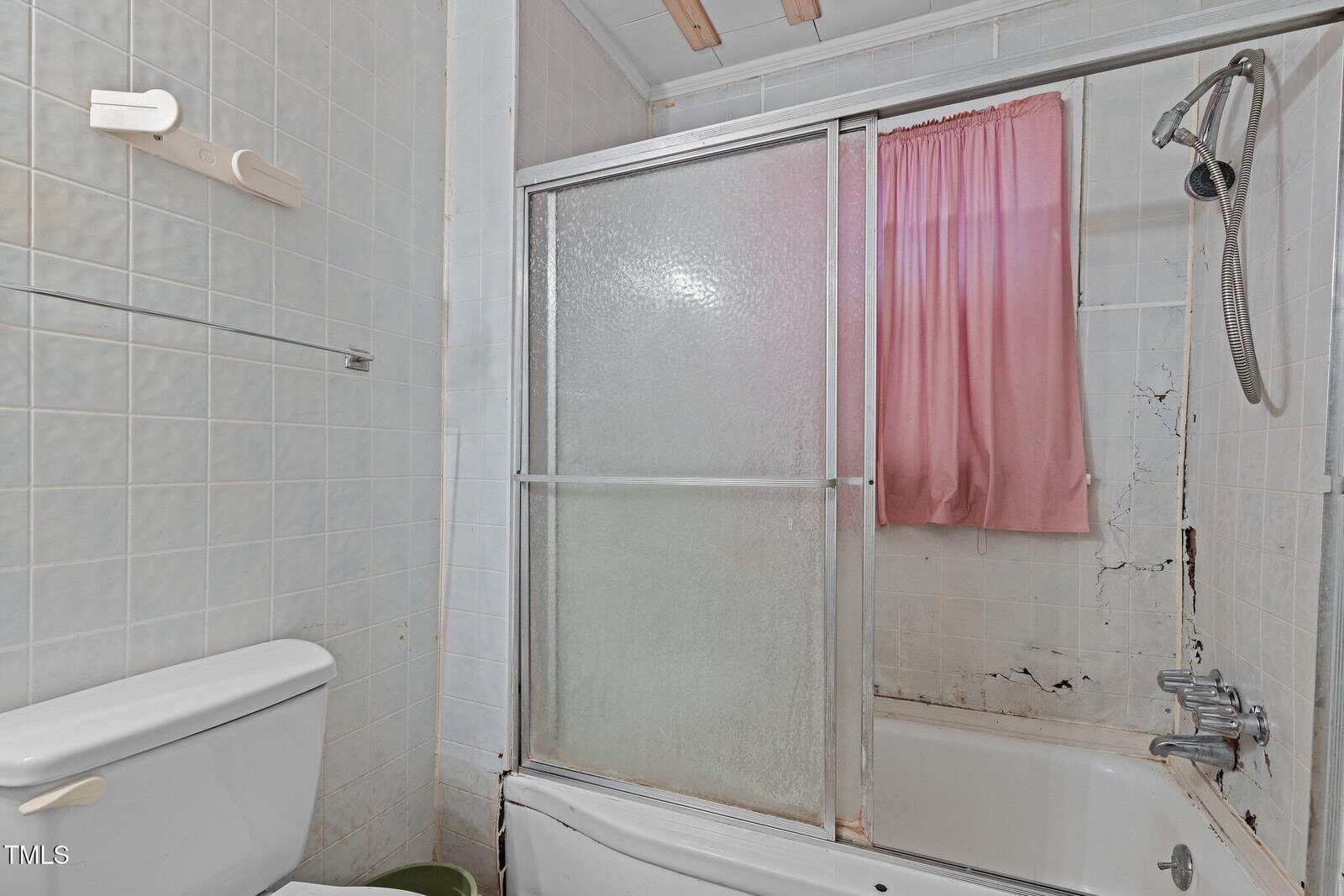 A bathroom with a white toilet, a green trash can, a tub with sliding glass doors, a handheld showerhead, pink curtain, and visible black mold or damage on tiles around the tub.
