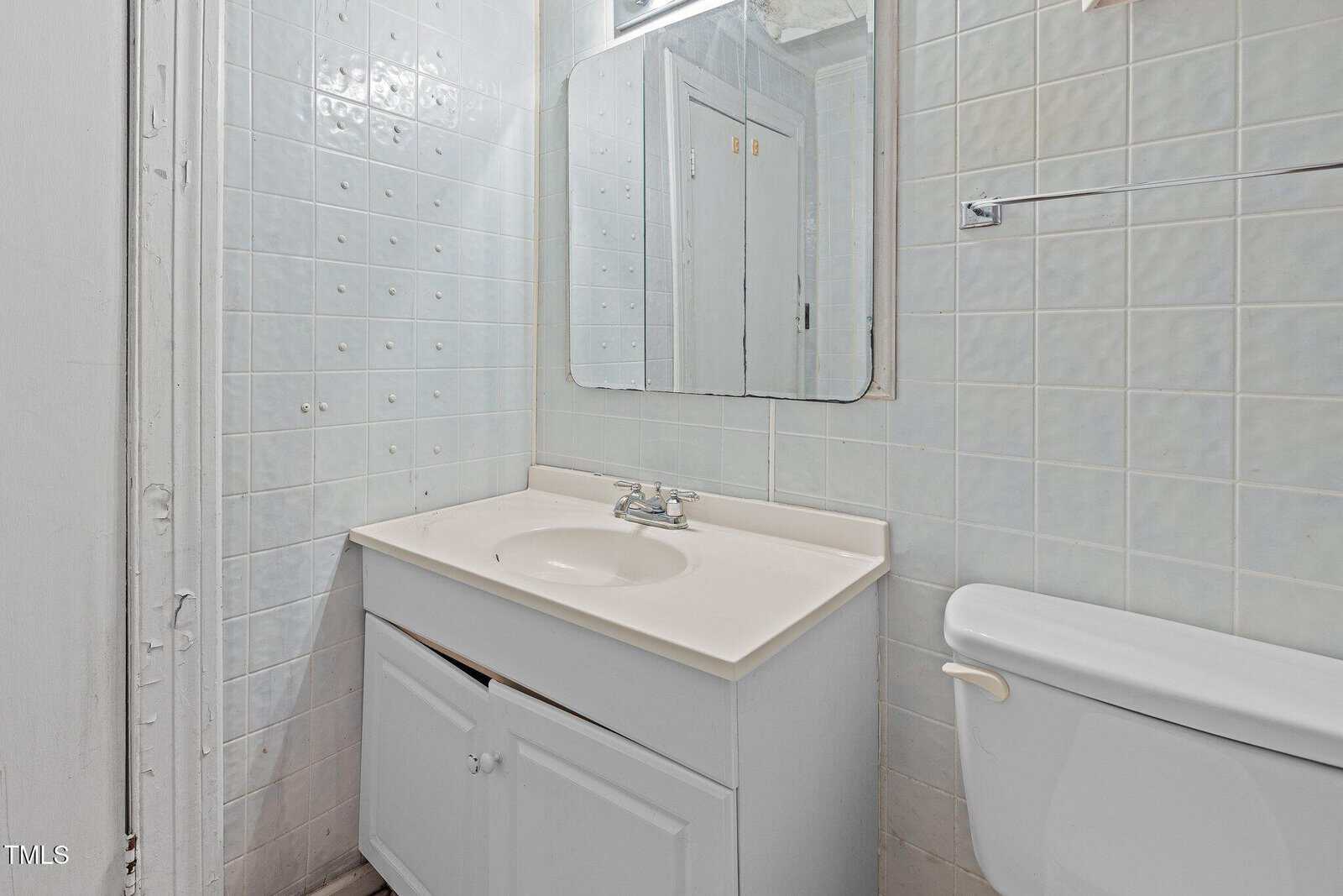 Small bathroom with white tiled walls, a white vanity with sink, a wall-mounted mirror above it, and a white toilet to the right. The space appears clean but minimally decorated.