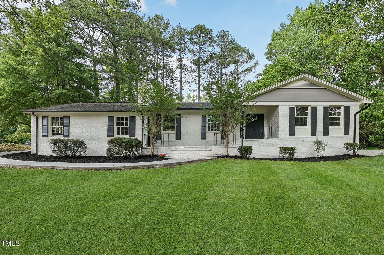 A single-story white brick house with black shutters and trim, set on a well-manicured green lawn, surrounded by tall trees and landscaping with bushes in front. Steps lead to a covered front entry.