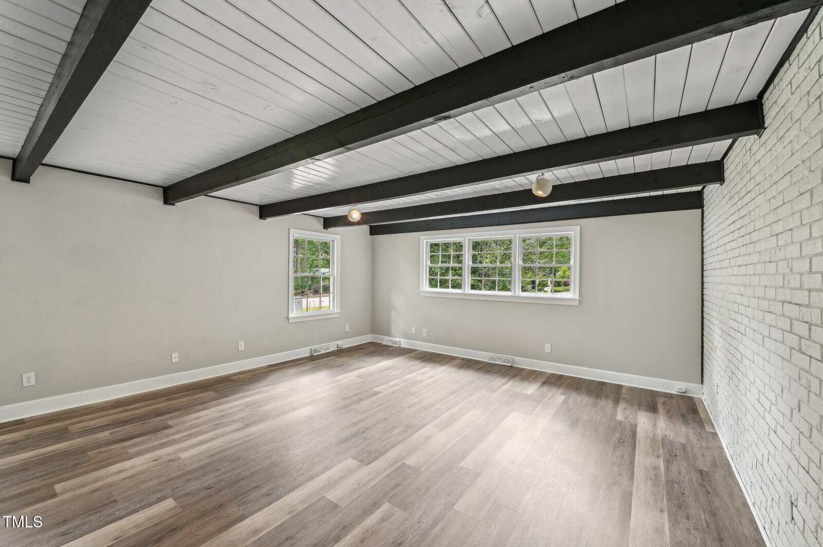 Spacious empty room with light wood flooring, white painted brick wall, large windows, and exposed black ceiling beams against a white ceiling. Walls are light gray. Natural light fills the space from the windows.
