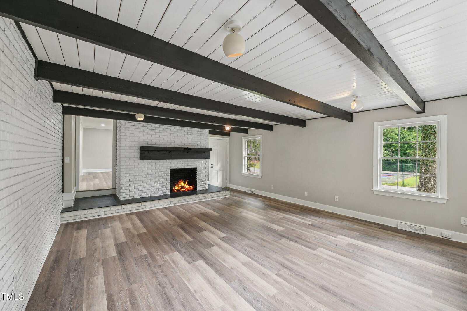 Spacious living room with white brick fireplace, exposed black ceiling beams, light gray walls, and wood-look flooring. Large windows let in natural light, and a doorway leads to another room. Fire is lit in the fireplace.
