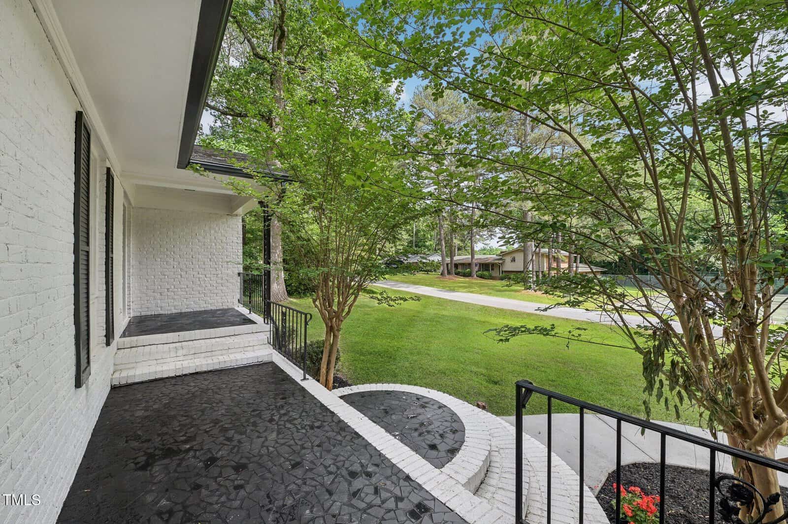 View from a covered front porch with black tiled flooring, white painted brick steps and railing, overlooking a green lawn, trees, and a quiet suburban street with houses in the background.