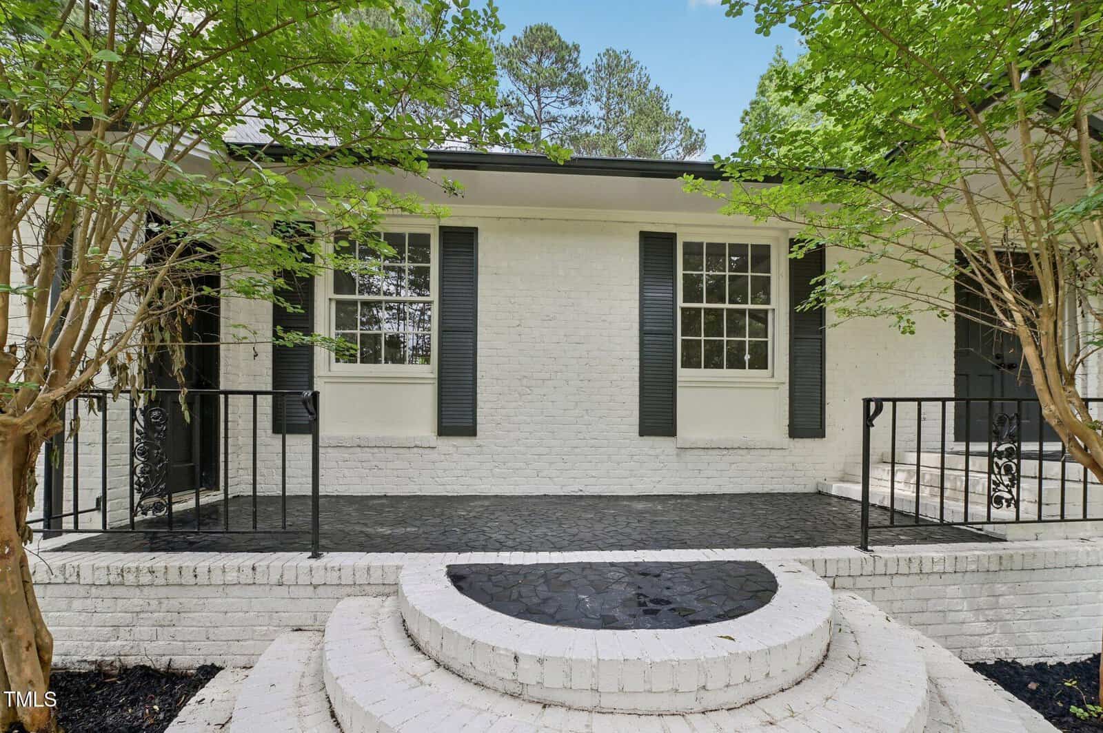 A white brick house with black shutters, a black front door, and a black-tiled porch. Two trees frame the entrance, and a short set of steps leads to the raised porch area with black metal railings.