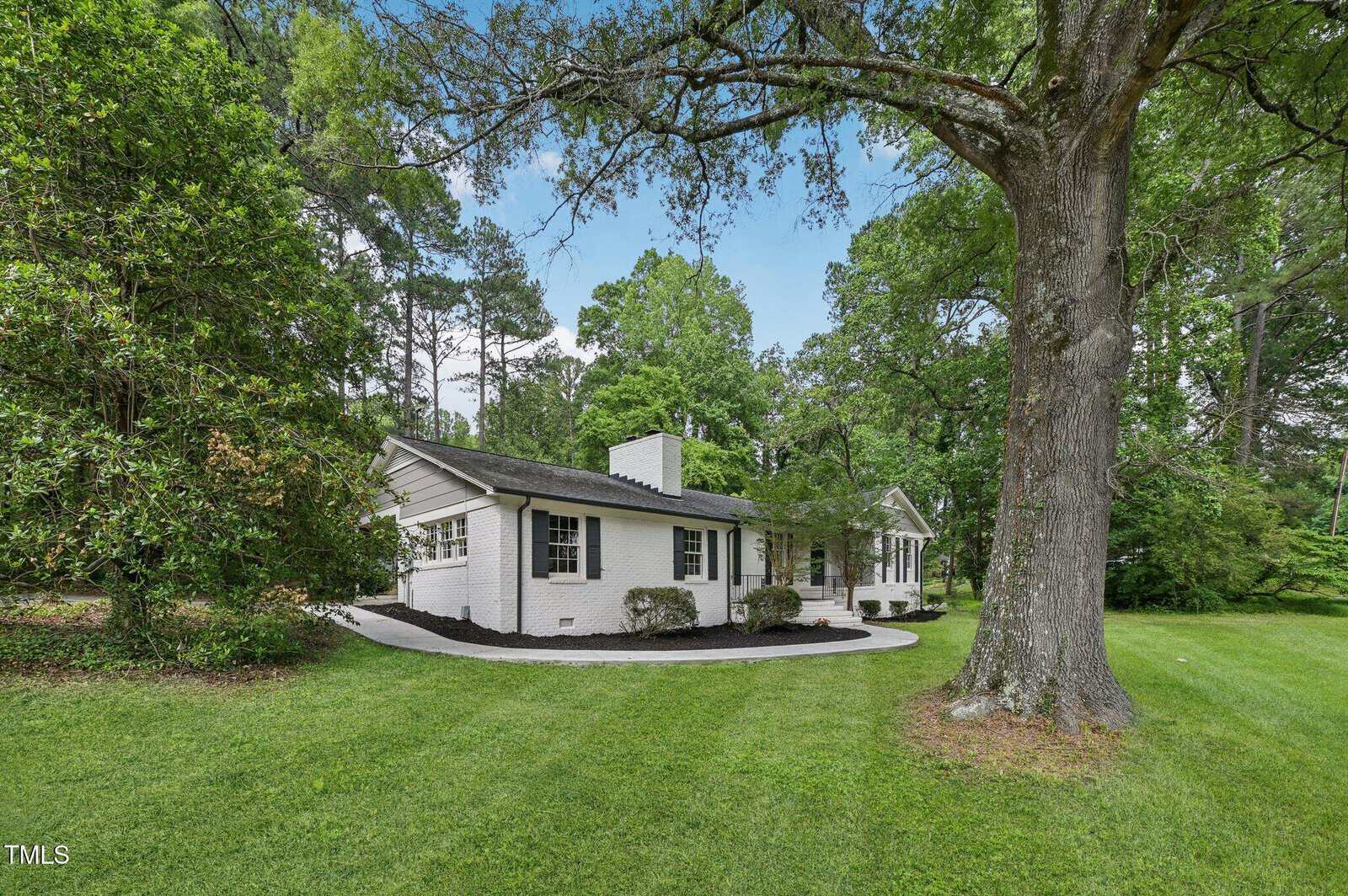 A white single-story house with a chimney sits among tall trees and lush green grass, with a curved pathway leading to the front door on a sunny day.