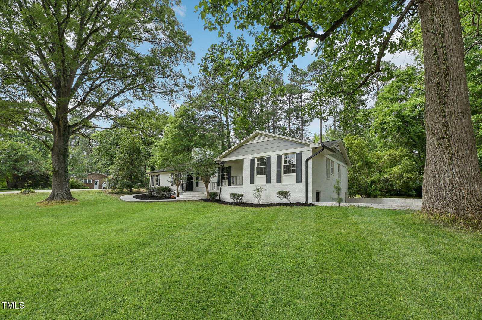 Single-story white brick house with black shutters and a gray roof, surrounded by lush green grass, large trees, and landscaping under a partly cloudy sky.