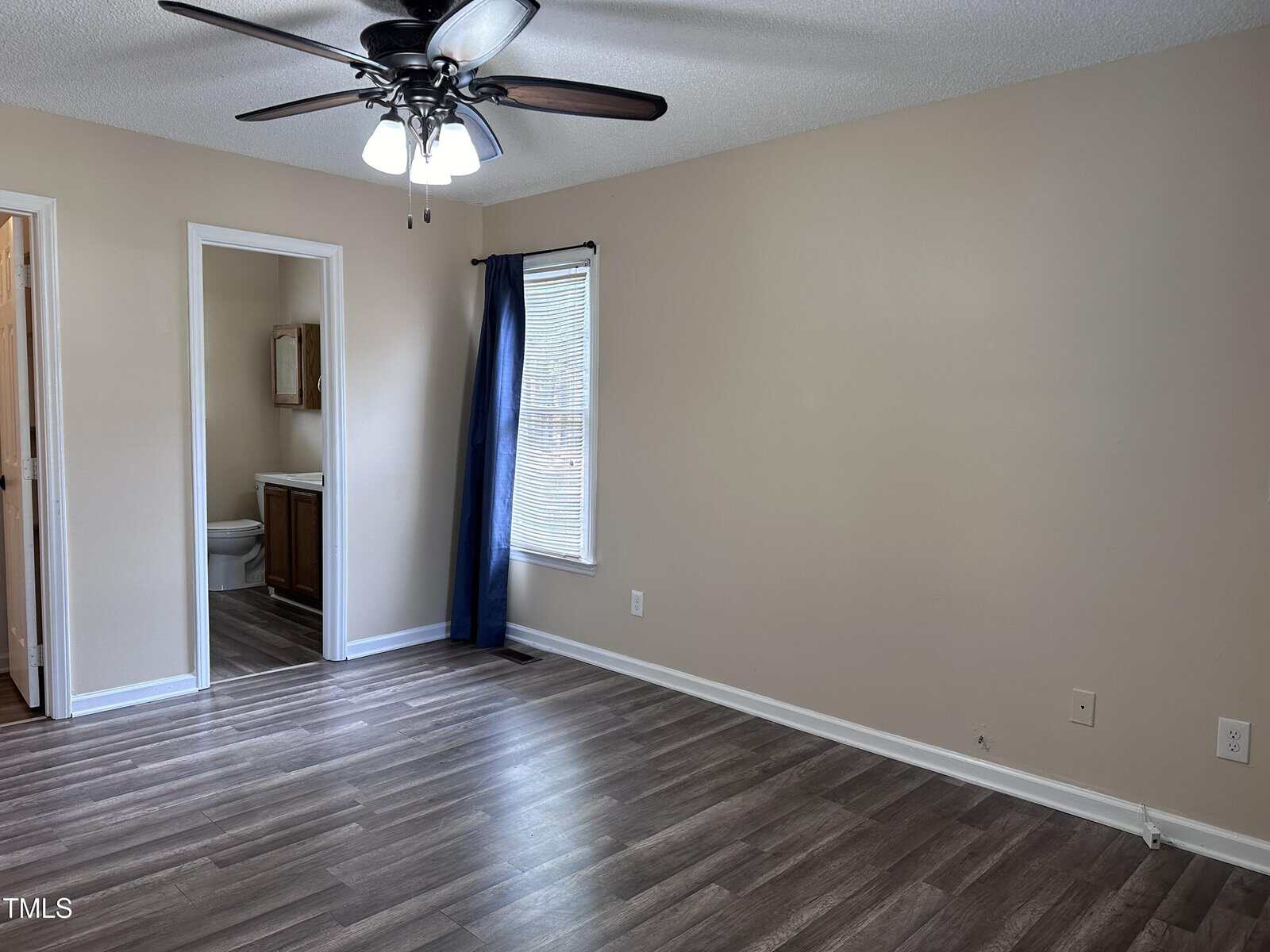 Empty room with beige walls, wood-look flooring, a ceiling fan with lights, a window with a blue curtain, electrical outlets, and a visible bathroom with a sink and toilet through an open doorway.