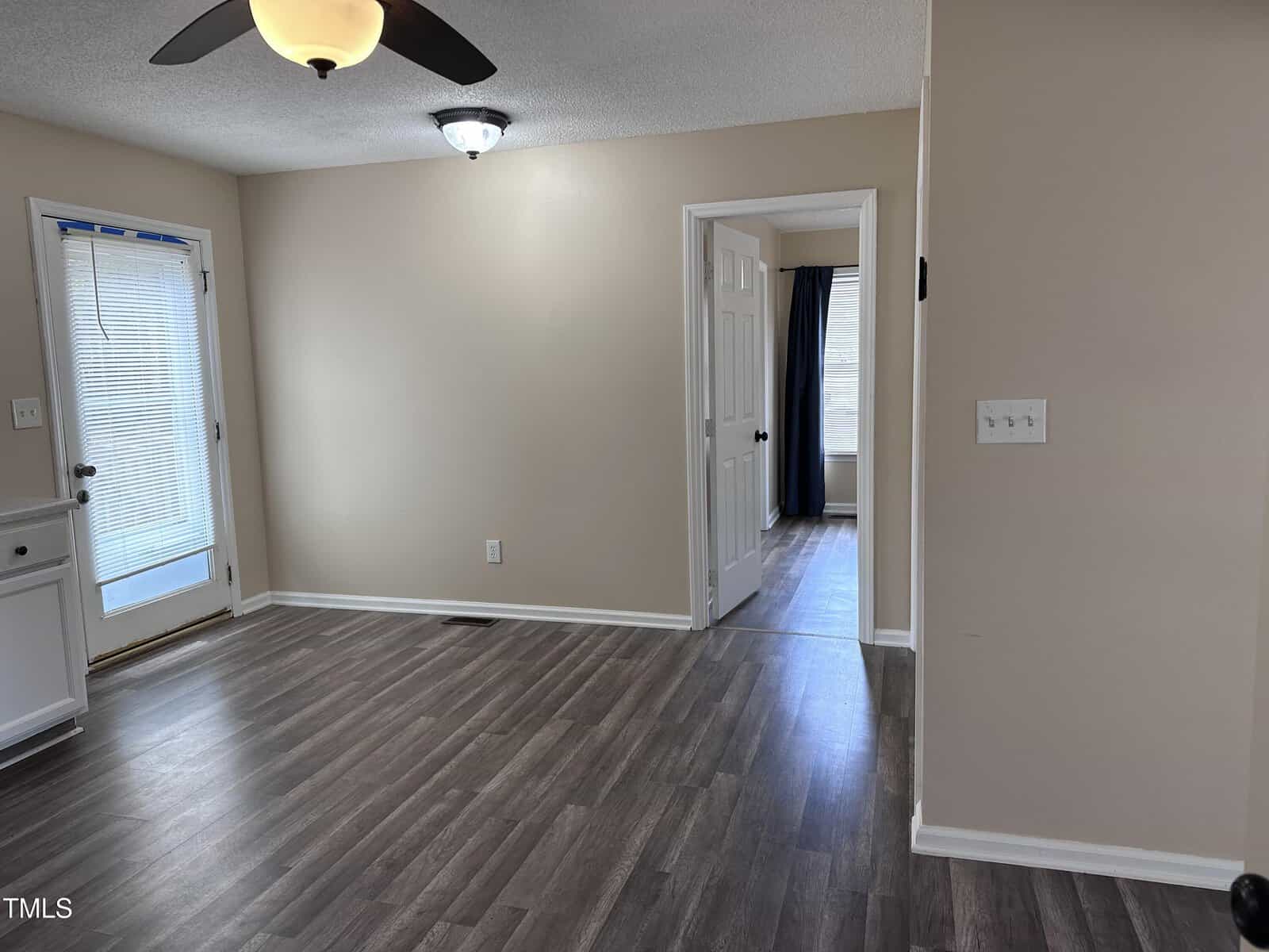 A room with light beige walls, wood-look flooring, a ceiling fan, and a door with blinds leading outside. An open doorway leads to another room with a window and dark curtains.