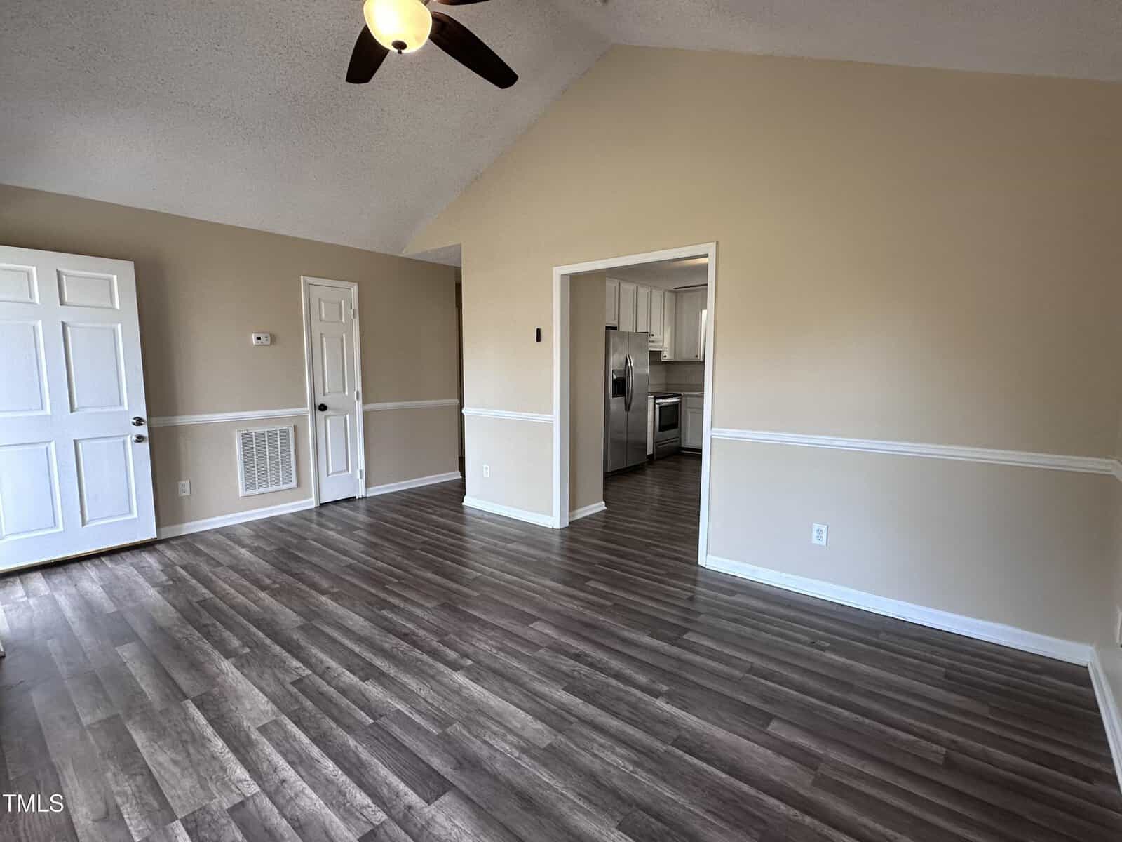 A spacious, empty room with vinyl wood flooring, beige walls, white trim, a ceiling fan, and two white doors. An entryway leads to a kitchen with white cabinets and stainless steel appliances.