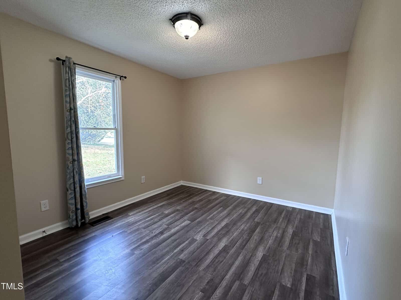 Empty room with beige walls, a single window with a blue curtain, a ceiling light fixture, and dark wood flooring. Sunlight is coming in through the window, and the room appears clean and unfurnished.