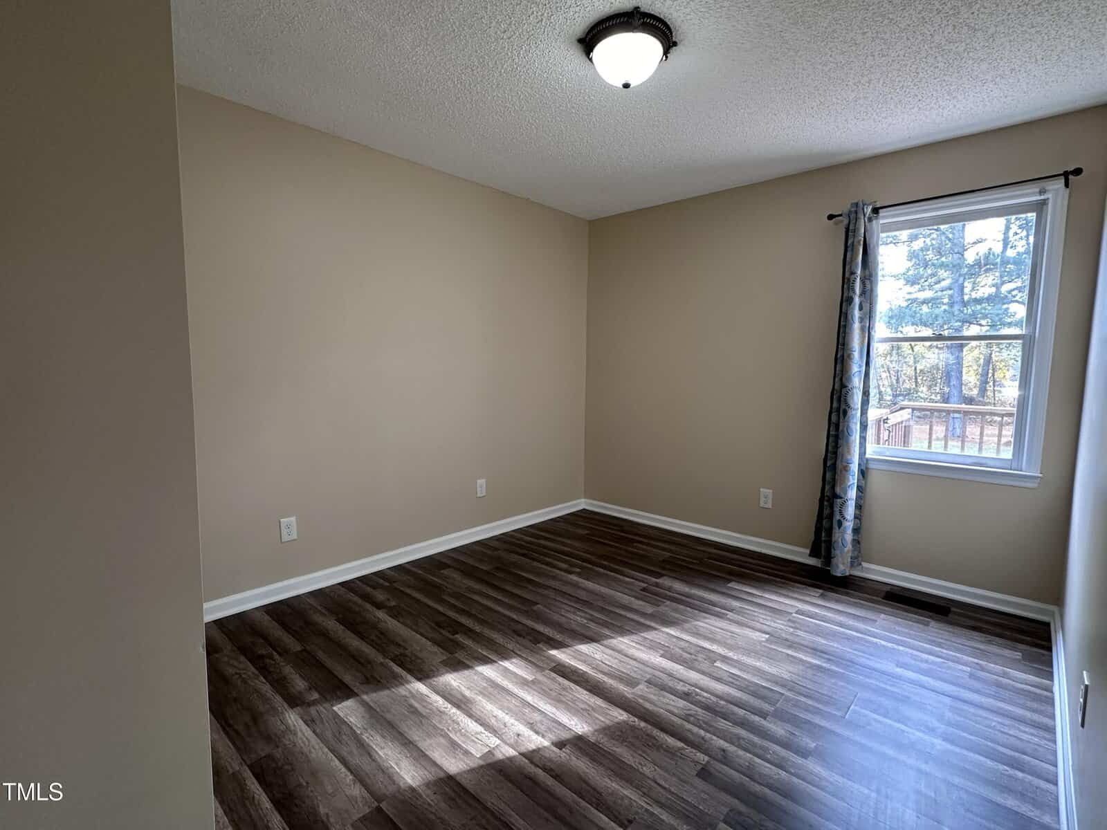 Empty room with beige walls, dark wood flooring, a ceiling light, a window with a curtain on the right wall, and visible electrical outlets along the walls. Sunlight is streaming in through the window onto the floor.