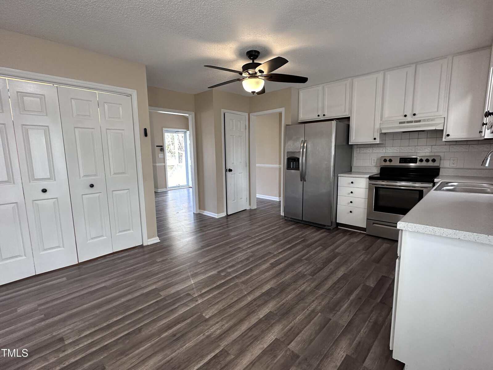 Modern kitchen with white cabinets, stainless steel appliances, a ceiling fan, double closet doors, and wood-style flooring. The space is well-lit with natural and ceiling light.
