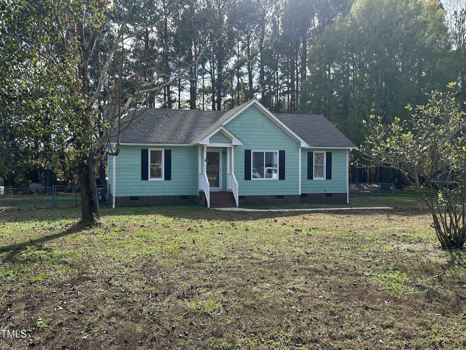 A small, light blue house with white trim and dark shutters sits on a grassy lot with scattered trees and a wooded background under a clear sky.