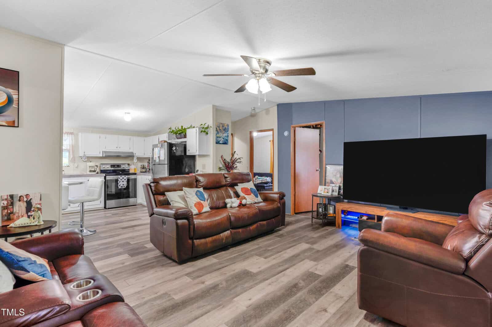 A living room with brown leather sofas, wood-look flooring, a ceiling fan with lights, a large flat-screen TV on a blue accent wall, and a view into a white kitchen with black appliances.