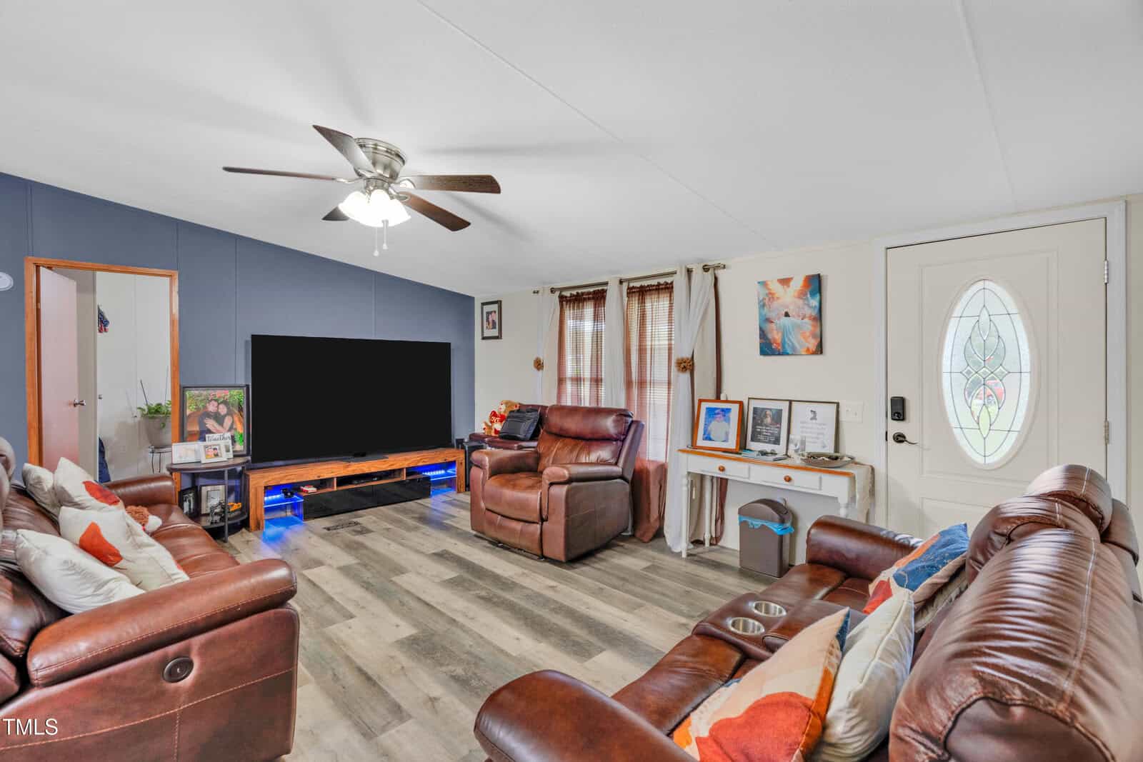 A living room with brown leather sofas, a large flat-screen TV on a wooden stand, wood-effect flooring, and a ceiling fan. A white door with a glass design and a small entry table are on the right side of the room.