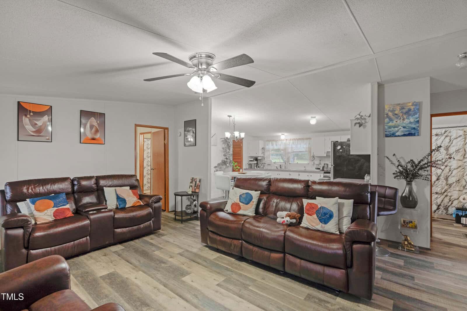 A living room with brown leather sofas, colorful accent pillows, wall art, and a ceiling fan. The room has wood-look flooring and opens to a kitchen and dining area with white cabinetry.