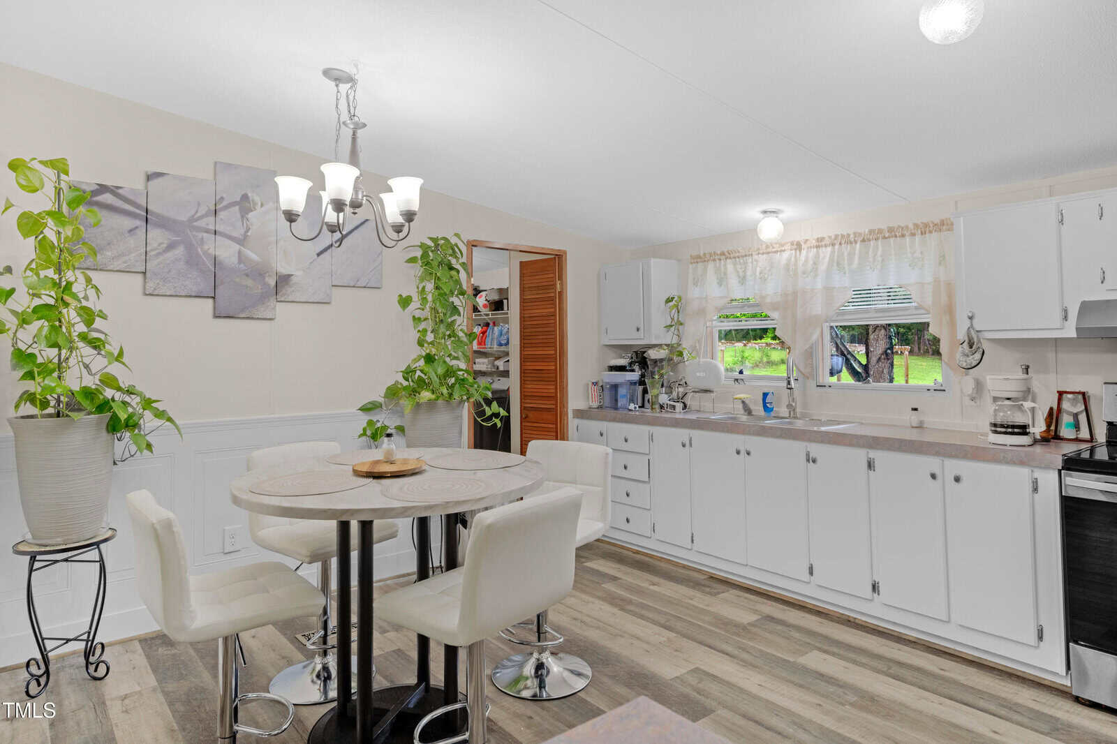 A kitchen and dining area with white cabinets, a round table with four white chairs, potted plants, and wood-patterned flooring. There are windows above the sink and wall decor above the table.