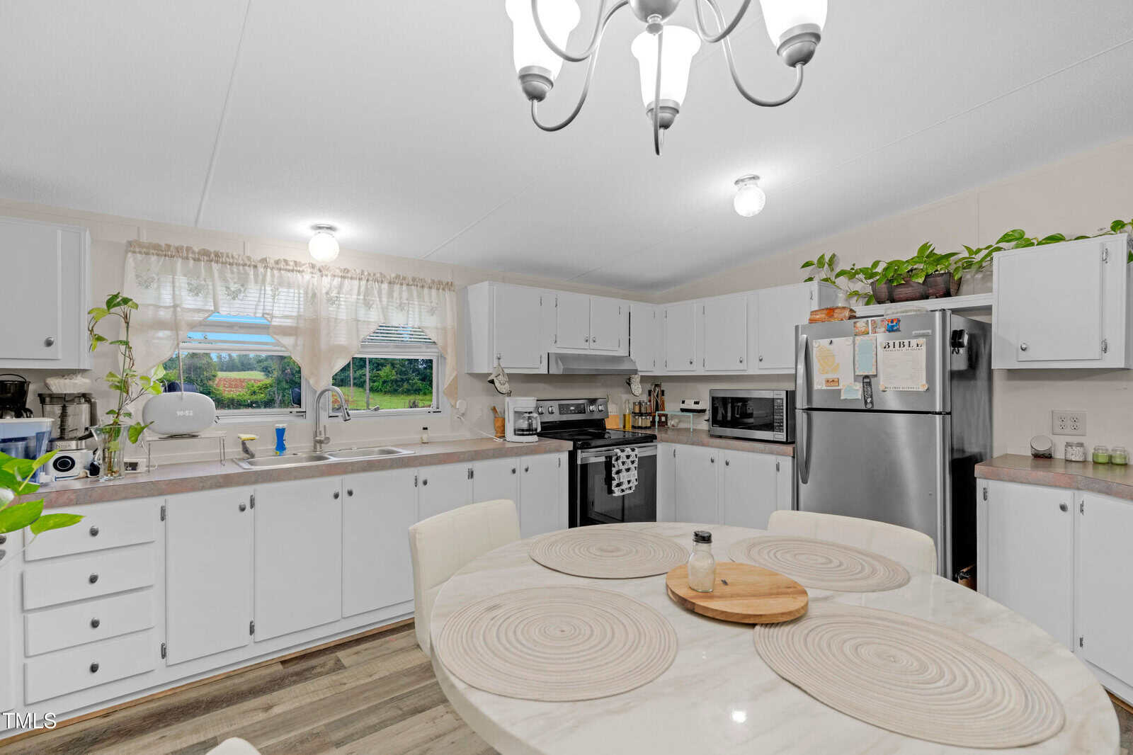 A bright kitchen with white cabinets, stainless steel appliances, a round table with placemats, and green plants on the counters and cabinets. Two windows above the sink let in natural light.