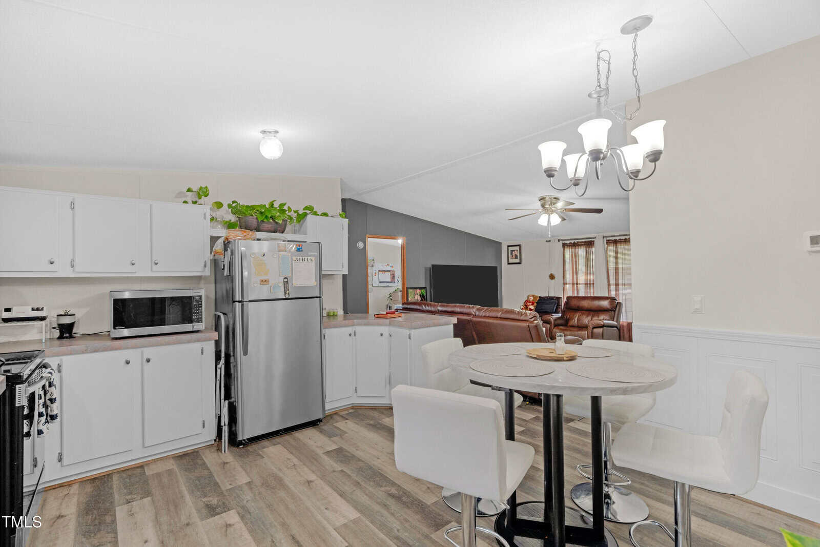 A modern kitchen with white cabinets, stainless steel appliances, a round dining table with four white chairs, wood-style flooring, and a living area with brown recliners and a large TV in the background.