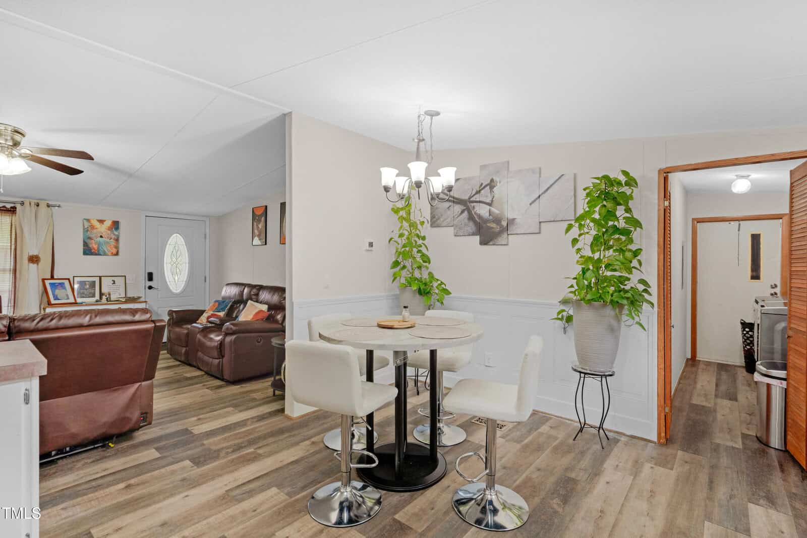A dining area with a round table and four white chairs is next to a living room with brown leather seating. There are two potted plants, wall art, and wood-style flooring throughout the space.