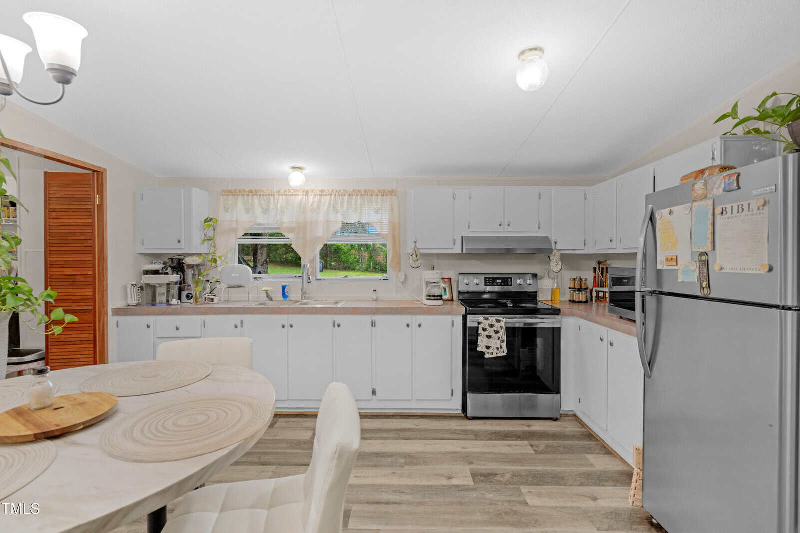 A bright kitchen with white cabinets, a black stove, a stainless steel refrigerator with magnets and papers, a round dining table with cushioned chairs, and light wood flooring. Two windows let in natural light.