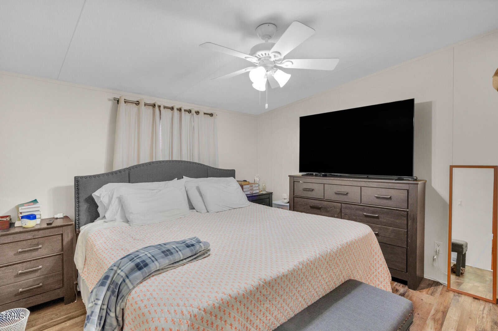 A bedroom with a large bed, gray headboard, and white bedding, flanked by two nightstands. A dresser with a flat-screen TV on top sits opposite the bed. A ceiling fan and a floor mirror are also visible.