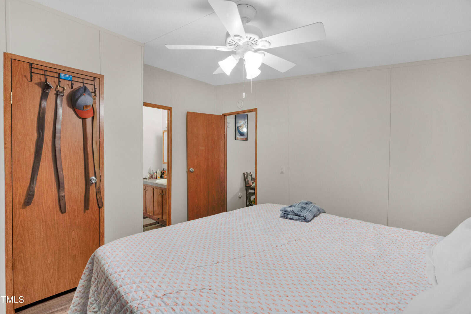 A bedroom with a ceiling fan, a bed with a patterned cover, a folded plaid shirt on the bed, and wooden doors—one with belts and hats hanging. An open door leads to a bathroom vanity.