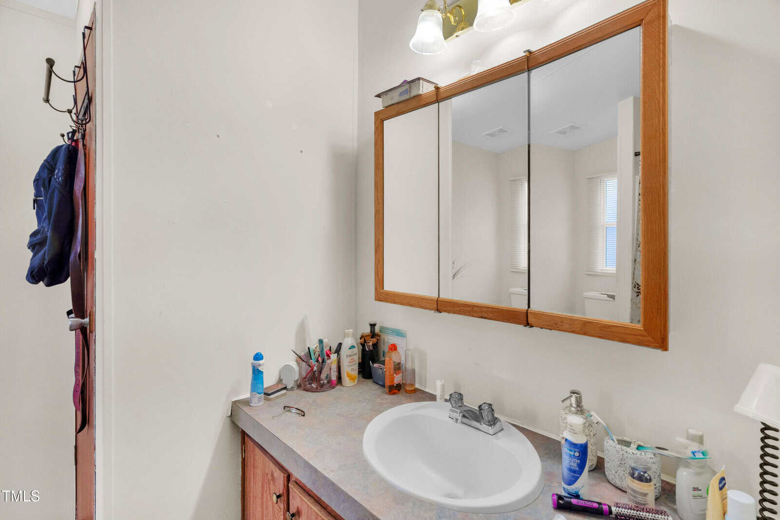 Bathroom countertop with various toiletries and beauty products around a sink, under a mirrored medicine cabinet. Overhead lights are on, and a towel is hanging on a door to the left.