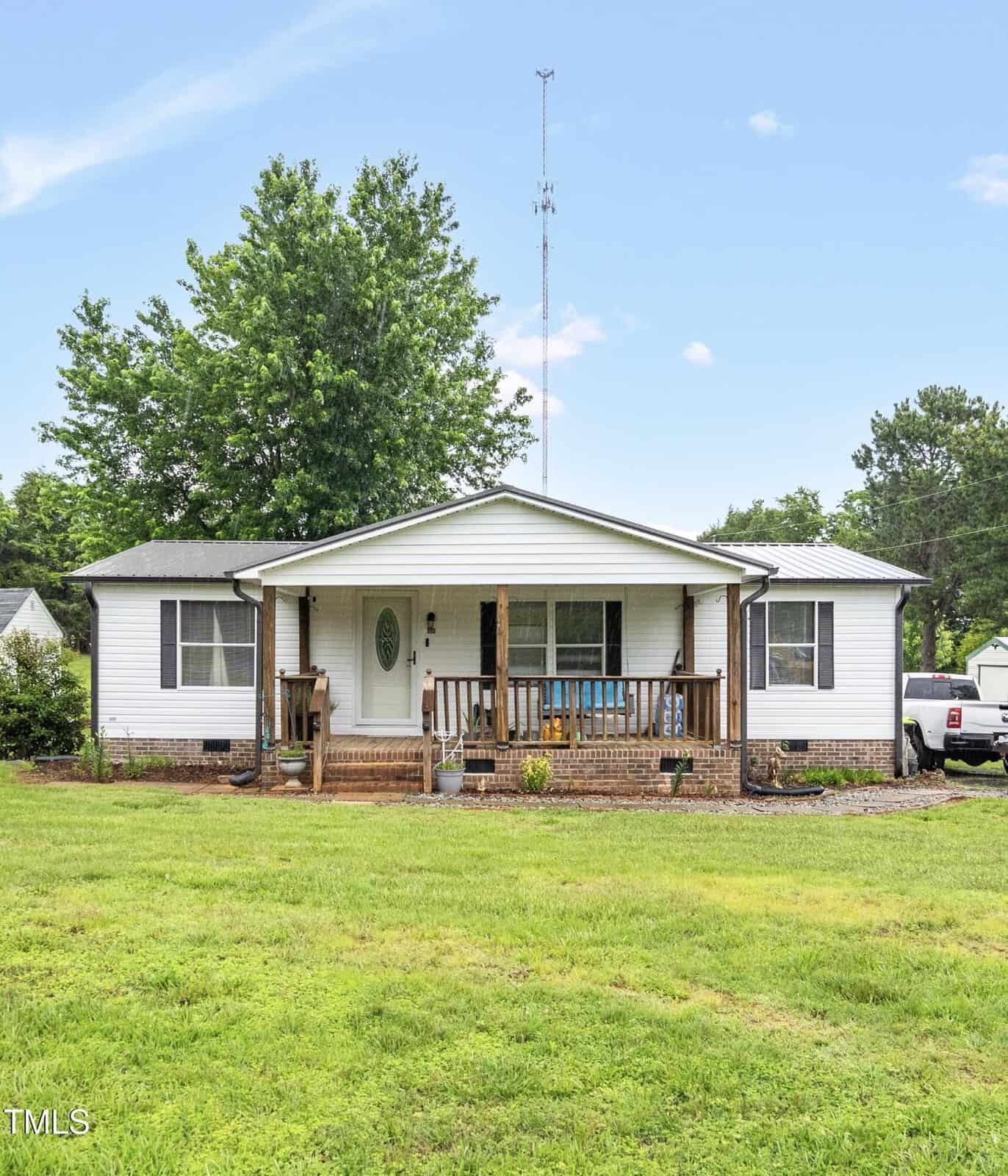 A single-story white house at 618 Puckett Street, Stovall, features a covered front porch with brick steps and wooden railing on a grassy lawn. A tree stands to the left, a truck is parked right, and a tall antenna rises behind the home.