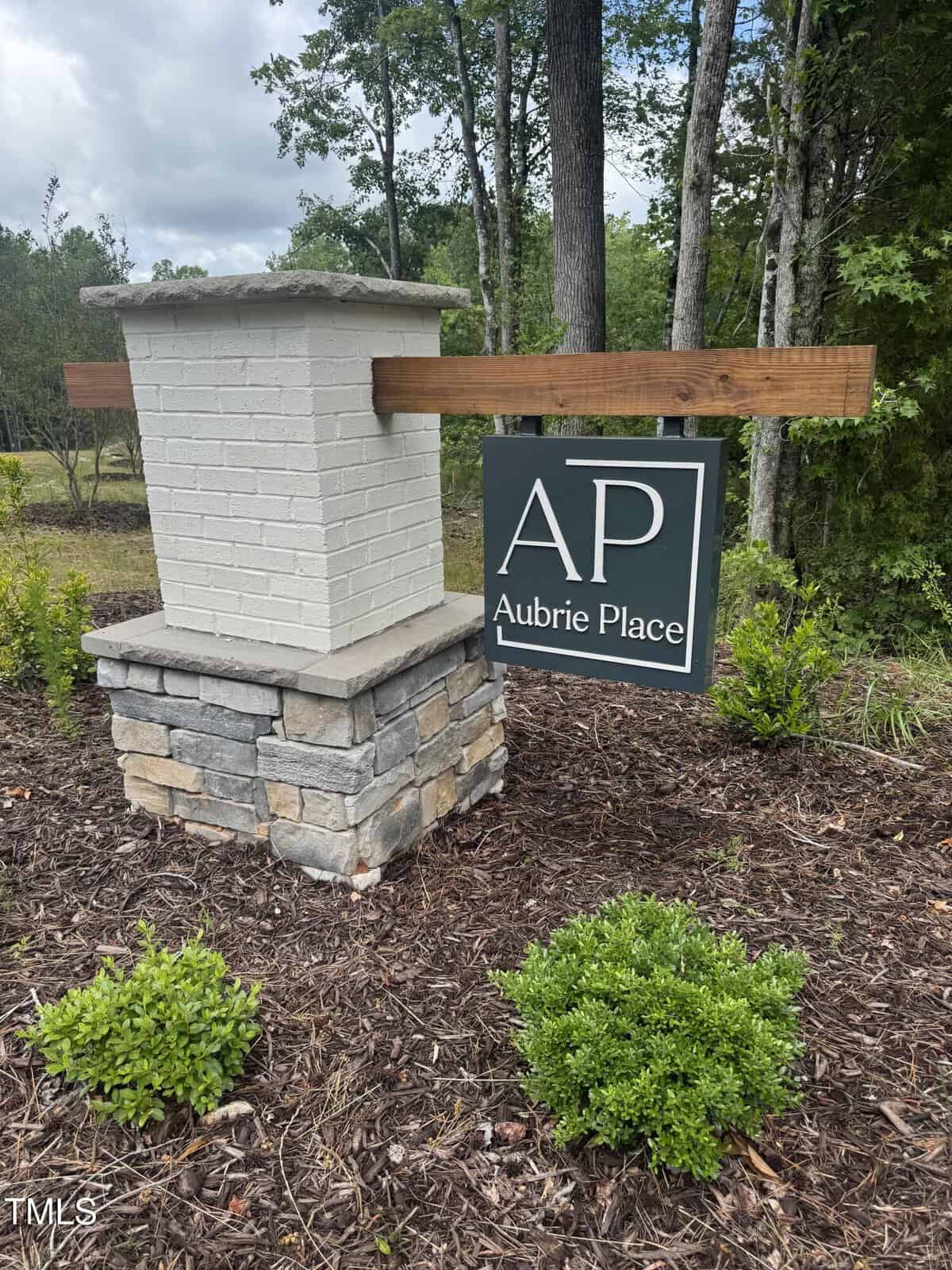 A rectangular sign reading "AP Aubrie Place" hangs from a wooden beam attached to a brick and stone pillar, set in landscaped mulch with small green shrubs, at 3801 Bennett Grove Drive in Wake Forest, with trees in the background.