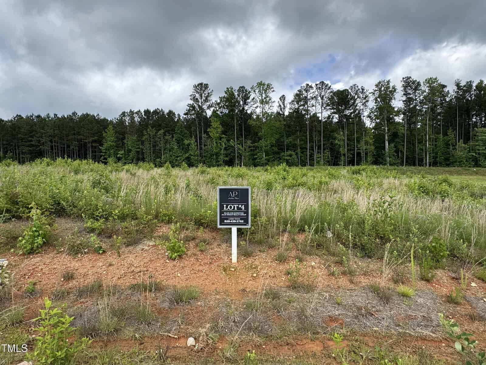 A grassy, undeveloped plot of land with sparse vegetation and a mix of dirt and grass. A sign in the center reads "Lot 14." Dense trees form a wooded background under a cloudy sky.