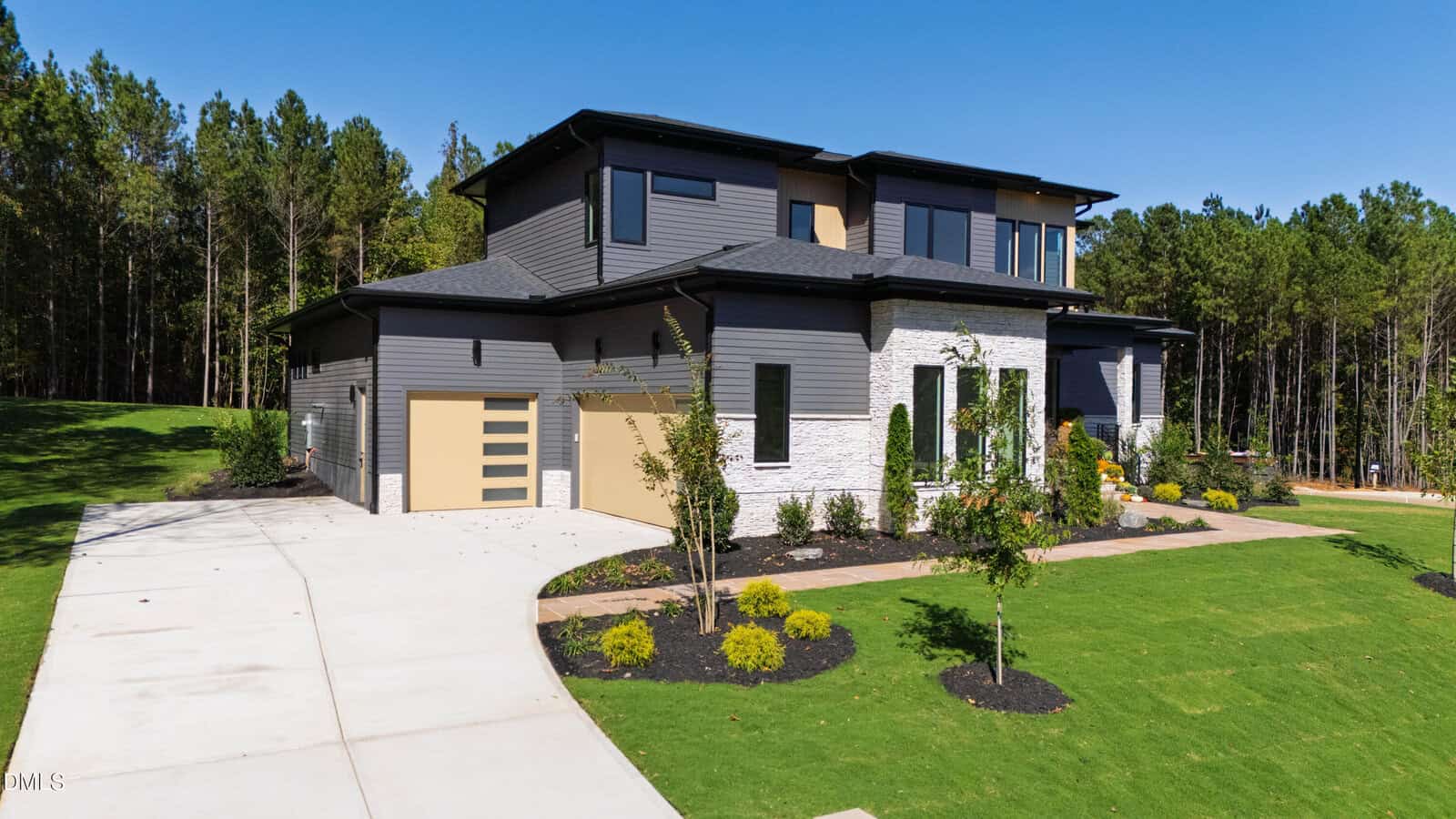 Two-story modern house with gray siding, white stone accents, and large windows. A curved driveway leads to a garage with a tan door. The lawn is landscaped with young trees and mulch beds. Dense woods border the property.
