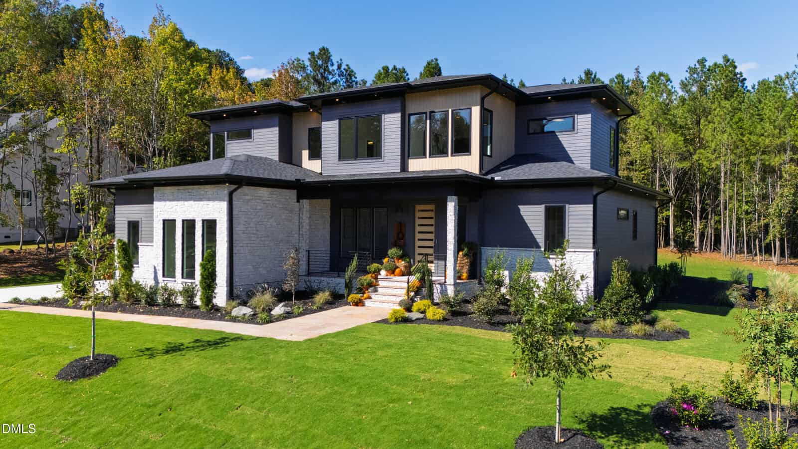 Modern two-story house with gray brick and siding exterior, large windows, a covered front porch with steps, landscaped yard, and surrounded by green lawn and trees under a clear blue sky.