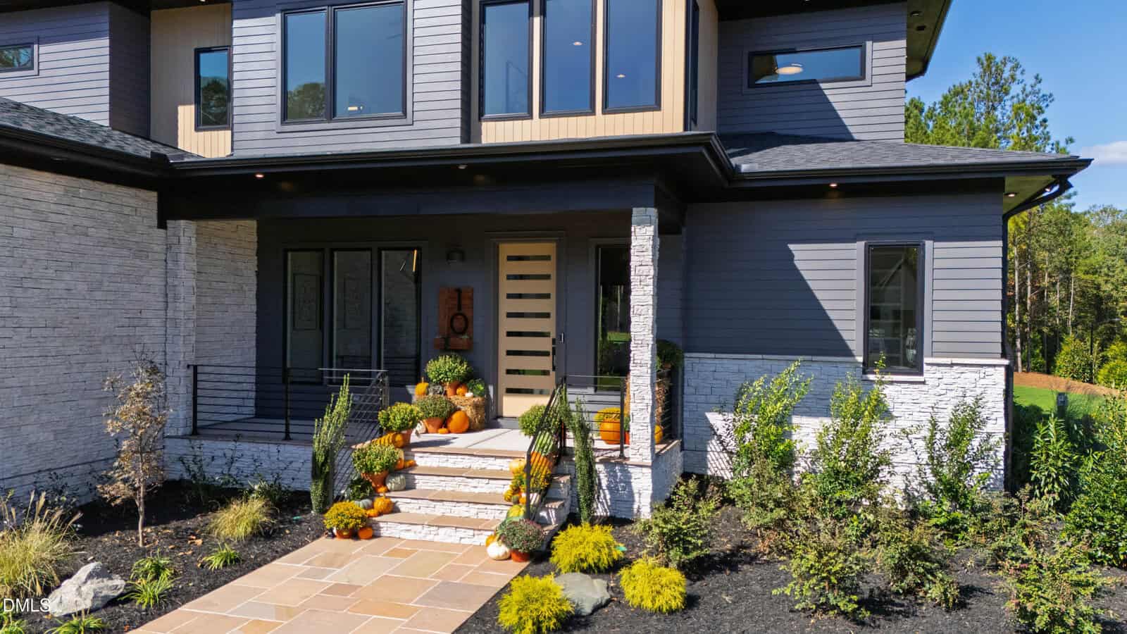 Modern two-story house with gray siding and white stone accents. The front porch is decorated with potted plants and orange pumpkins, while neatly trimmed bushes line the walkway and stairs leading to the entrance.