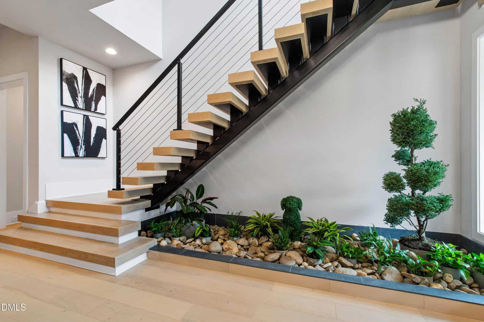 Modern staircase with light wood steps and black railing above an indoor garden bed containing rocks, green plants, and topiary, set against a white and gray wall with abstract artwork on the left.