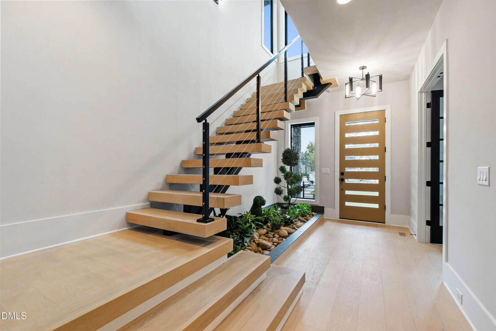 Modern entryway with floating wooden stairs, glass railing, light wood floors, and a landscaped indoor planter with rocks and plants underneath the stairs. A contemporary front door with horizontal glass panels is visible.