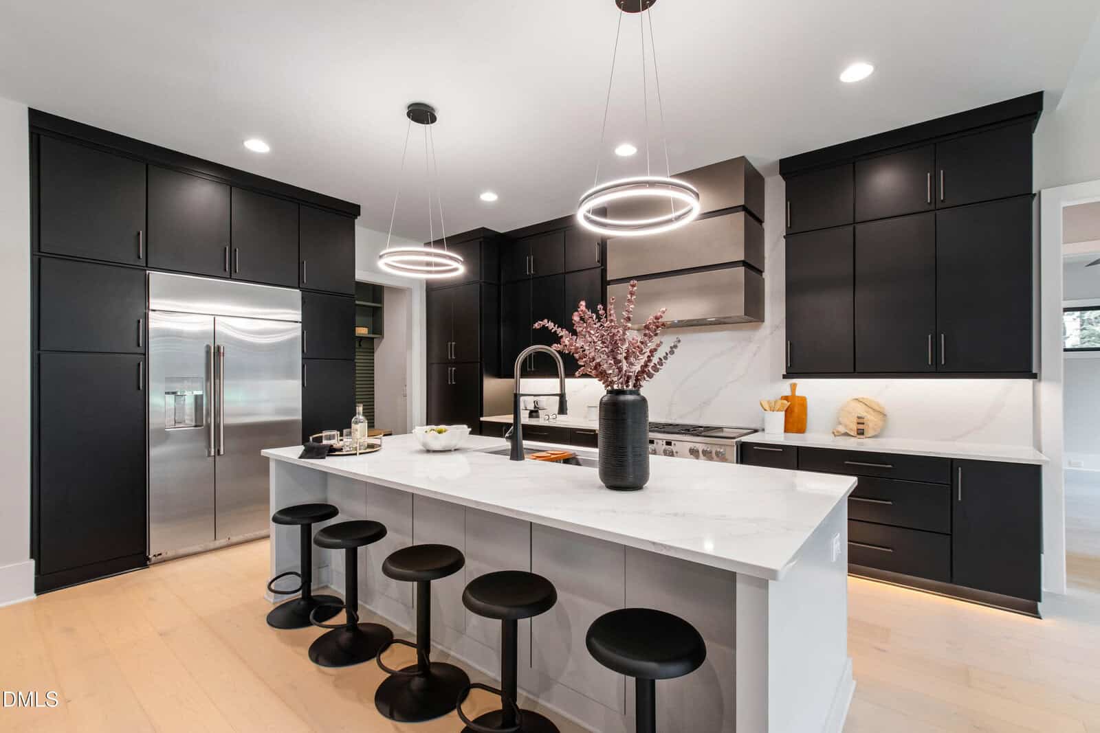 Modern kitchen with black cabinets, stainless steel appliances, a large island with four black stools, two circular pendant lights, and a white countertop featuring a black vase with pink branches.