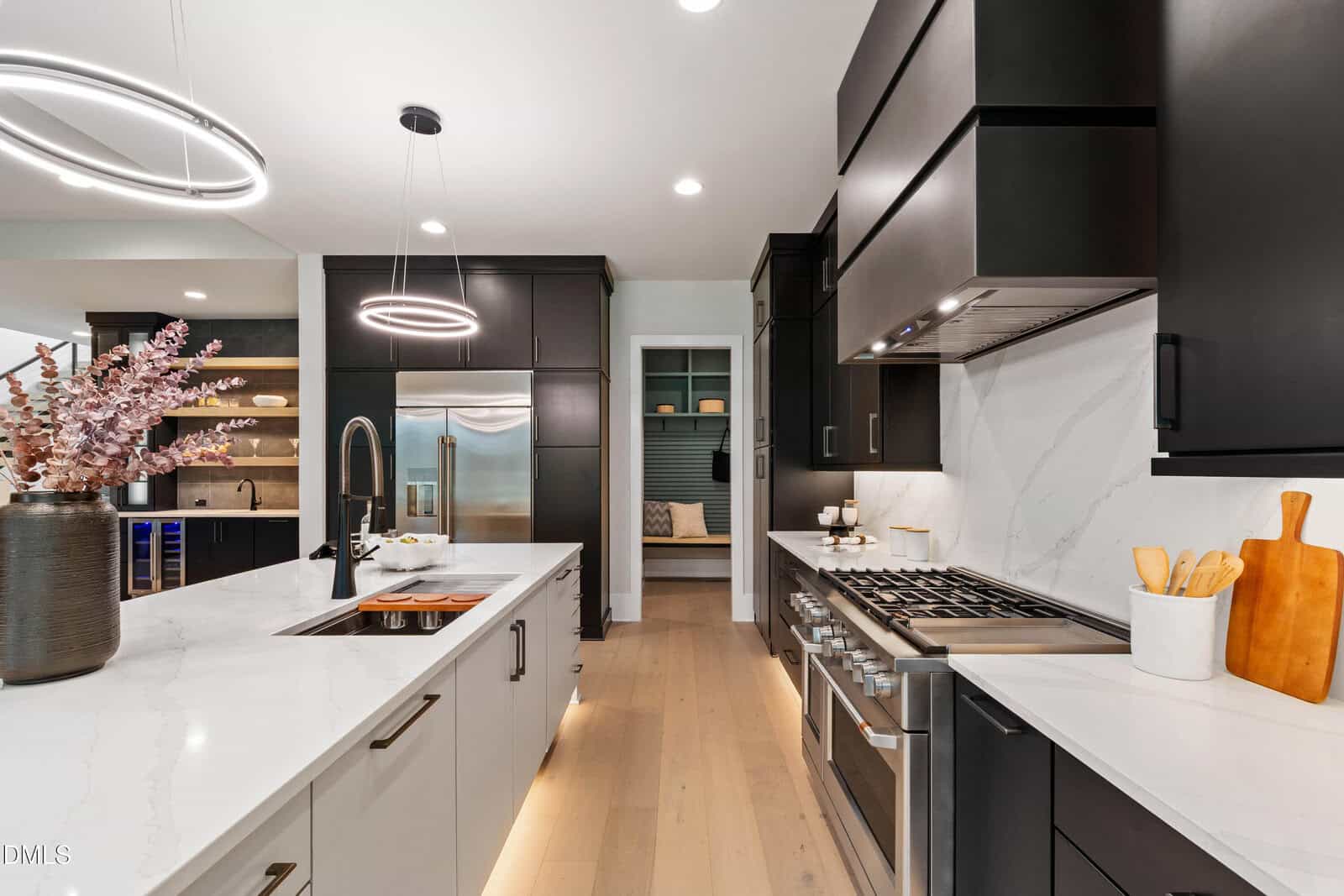 Modern kitchen with black and white cabinetry, stainless steel appliances, a gas stove, a large island with a sink, light wood floors, and minimalist lighting. Pink flowers in a vase sit on the island.