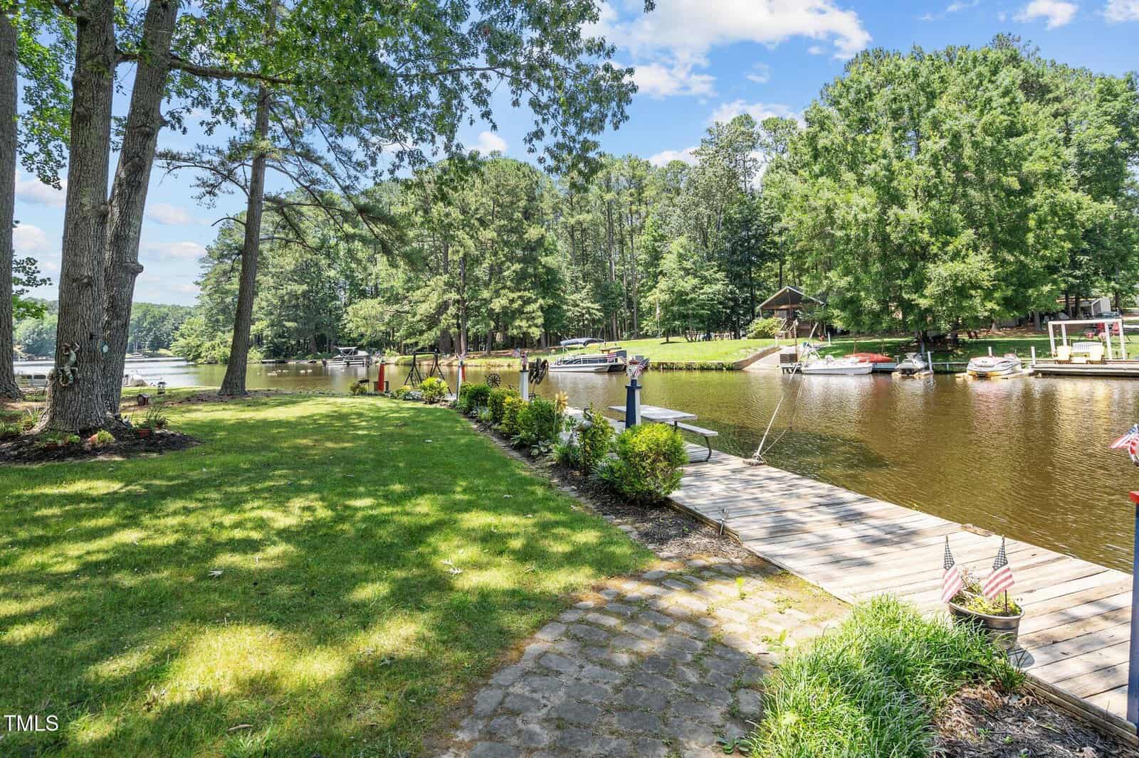 A grassy yard with a stone pathway leads to a wooden dock on a calm river. Several boats are moored along docks, and tall trees line both sides of the water under a partly cloudy sky.