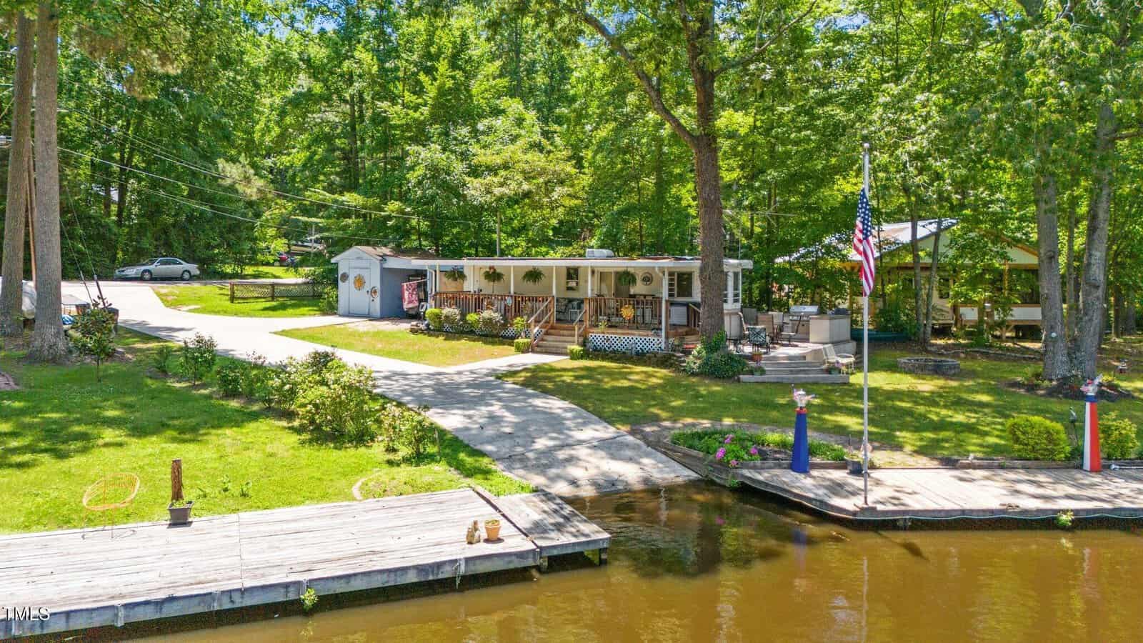 Single-story home with a covered porch sits by a lake, surrounded by trees and grassy yard. There are wooden docks, an American flag, and a parked car in the driveway. A small shed is visible near the house.
