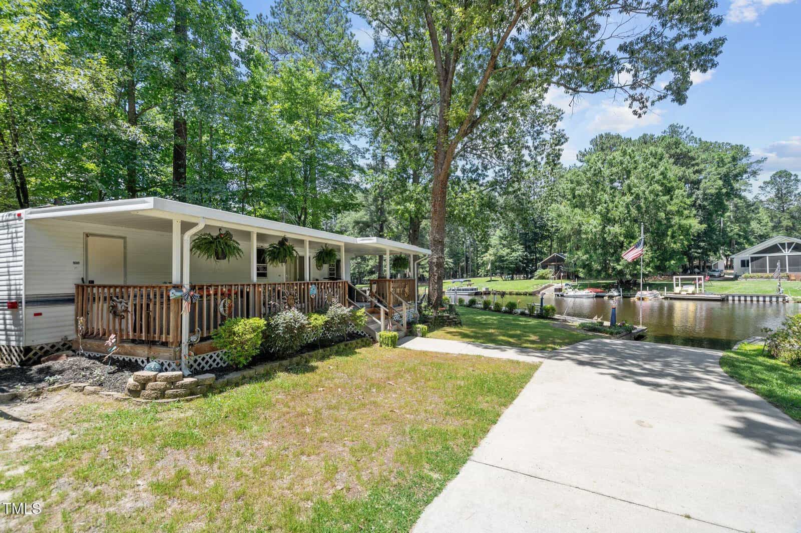 A charming white mobile home at 108 Big Horn Drive in Louisburg features a covered porch adorned with potted plants, set near a canal with a dock. Surrounded by trees and grass on a sunny day, the property includes a flag by the water and a paved driveway leading to the home.