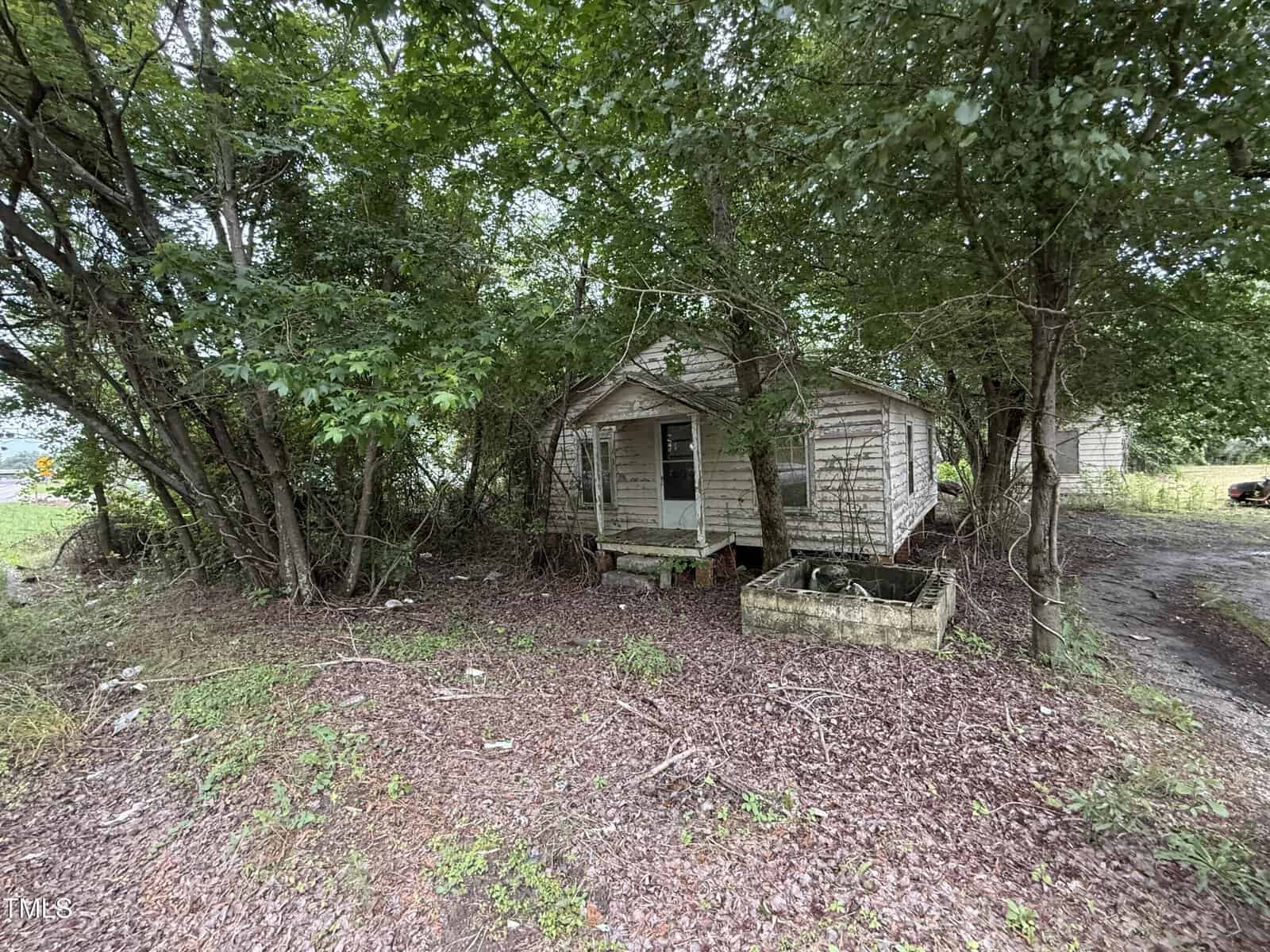 A small, weathered wooden house sits surrounded by overgrown trees and brush at 00 Warrenton Road. The house has peeling paint, a missing step at the entrance, and an empty, rectangular concrete planter in front.