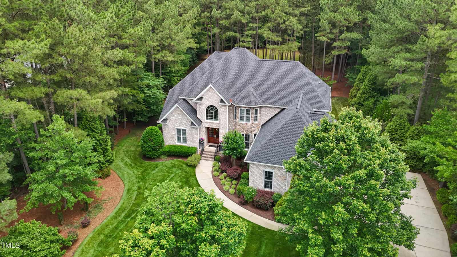 A two-story brick house with a dark gray roof is surrounded by green trees and landscaped lawns, with a curved walkway leading to the front entrance and a driveway on the right.