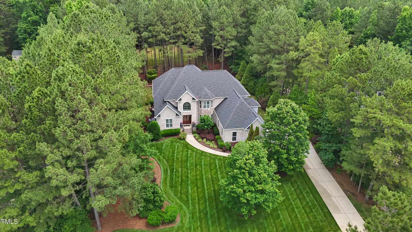 Aerial view of a large two-story house with a gray roof, surrounded by trees and greenery, featuring a curved driveway, landscaped lawn, and a wooded backdrop.