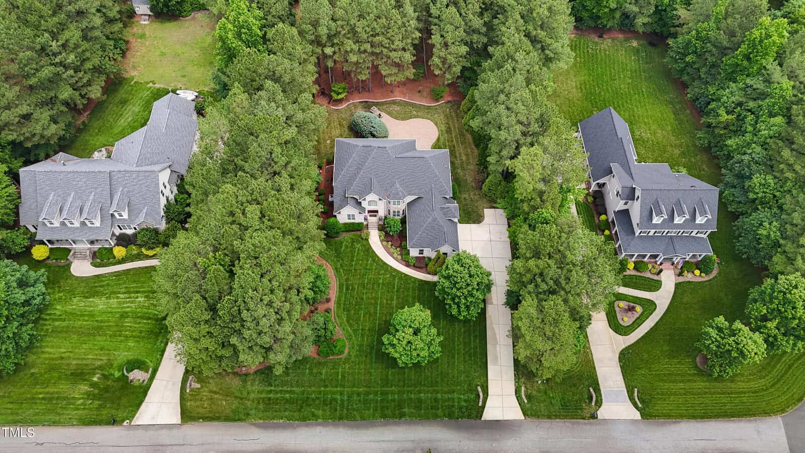 Aerial view of three large suburban houses with grey roofs, surrounded by green lawns, trees, and landscaped gardens, each with a driveway leading to the street at the bottom of the image.