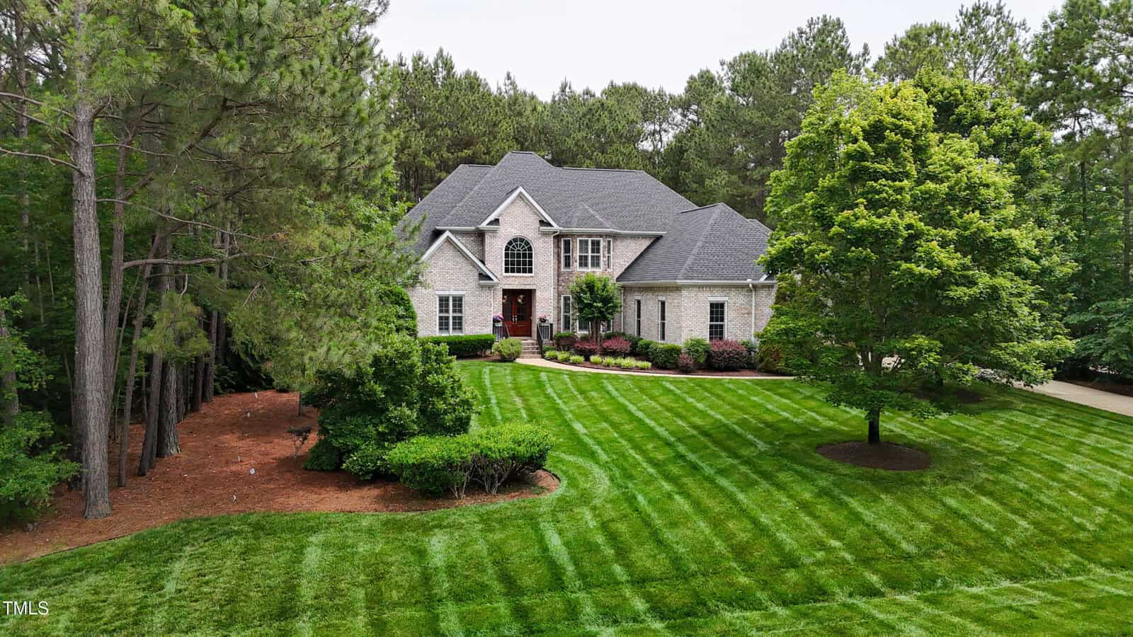 A two-story house with a light brick exterior sits amid a large, well-manicured lawn at 3004 Krogen Court in Creedmoor, featuring freshly mowed stripes and surrounded by trees and landscaping under an overcast sky.
