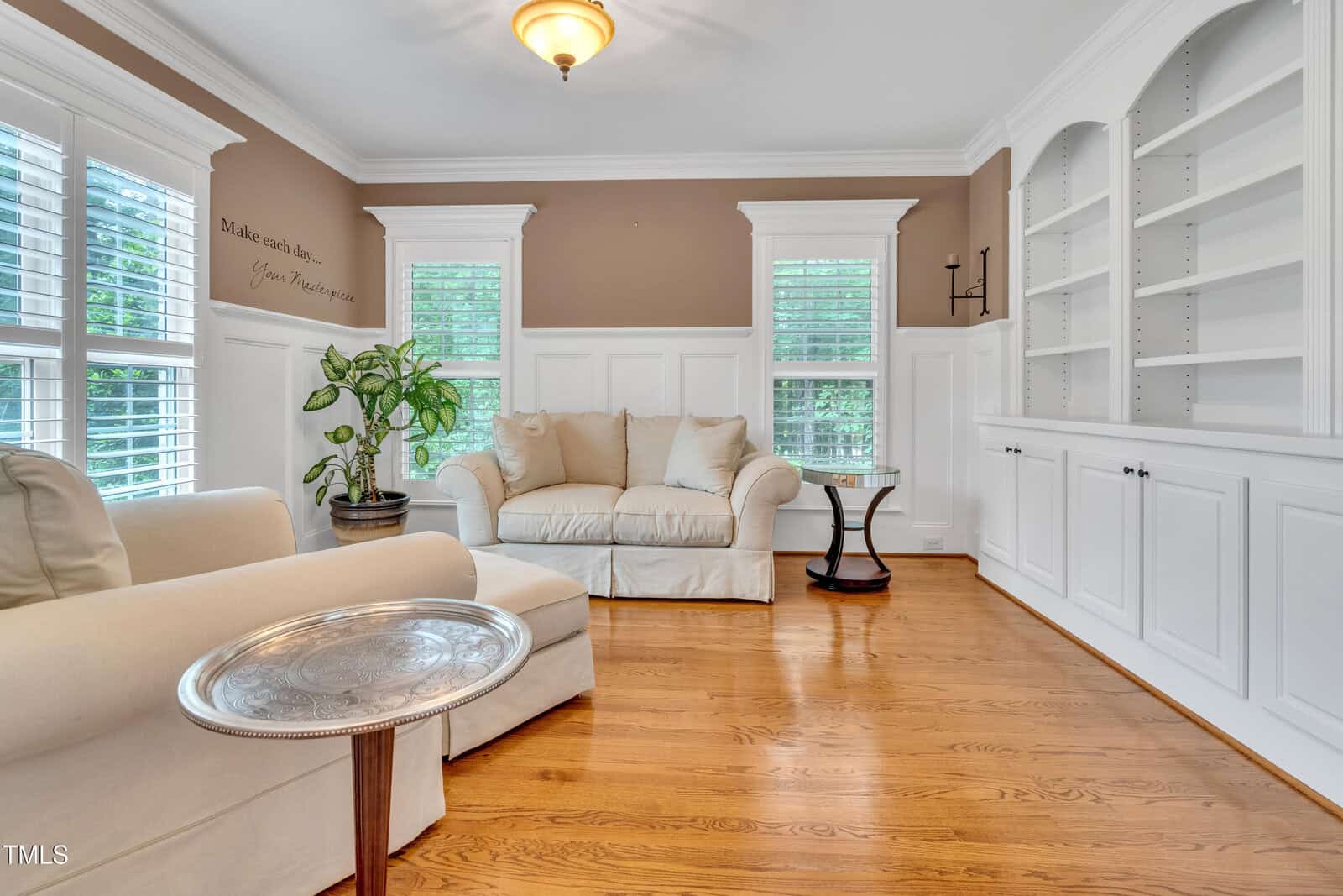 Bright living room with hardwood floors, white built-in shelves, beige furniture, a green potted plant, and large windows with white shutters. A round silver side table is in front of a chaise lounge.